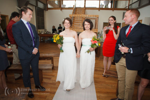 Two brides, both in white dresses, walk up their wedding aisle together holding bouquets