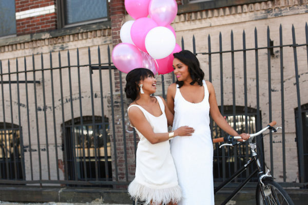 Two African-American brides walk down a city street in white dresses, carrying pink and white balloons. Photo by Amber Marlow Photography