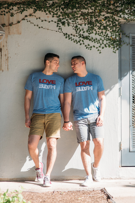 Two men stand under a bridge, holding hands. They both wear blue t-shirts with the words "Love Conquers Hate" written on them. - Photo by Elissa R Photography austin-gay-engagement-photos-08