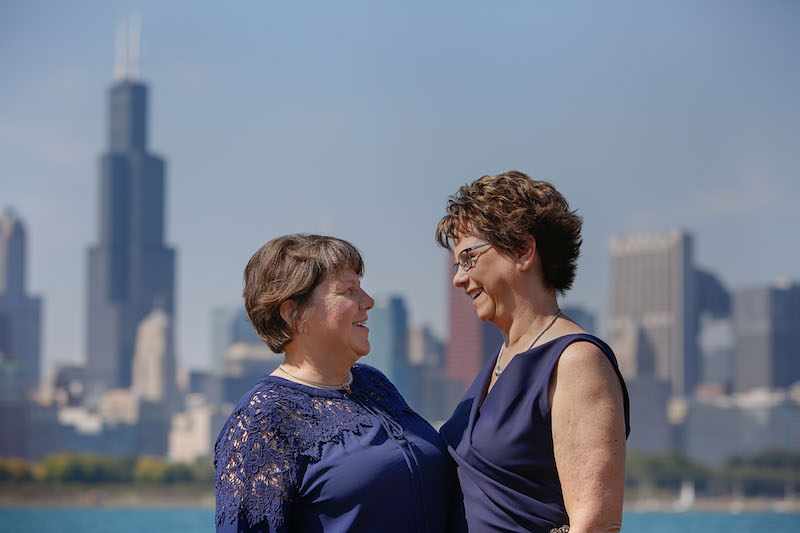 Pam, left, in a navy blouse; and Mary, right, in a navy sleeveless dress, stand looking into each other's eyes against the Chicago skyline and lakefront.