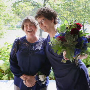 Pam, left, in a navy blue top and khaki trousers stands with Mary, right, in a navy blue wrap dress during their Chicago elopement. Pam wears a wrist corsage; Mary holds a colorful bouquet. Chicago Elopement Mary Pam Wedding 109 Evanston Ceremony