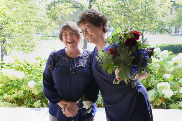 Pam, left, in a navy blue top and khaki trousers stands with Mary, right, in a navy blue wrap dress during their Chicago elopement. Pam wears a wrist corsage; Mary holds a colorful bouquet. Chicago Elopement Mary Pam Wedding 109 Evanston Ceremony