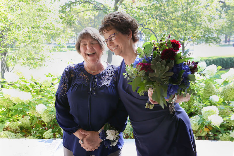 Pam, left, in a navy blue top and khaki trousers stands with Mary, right, in a navy blue wrap dress during their Chicago elopement. Pam wears a wrist corsage; Mary holds a colorful bouquet. Chicago Elopement Mary Pam Wedding 109 Evanston Ceremony