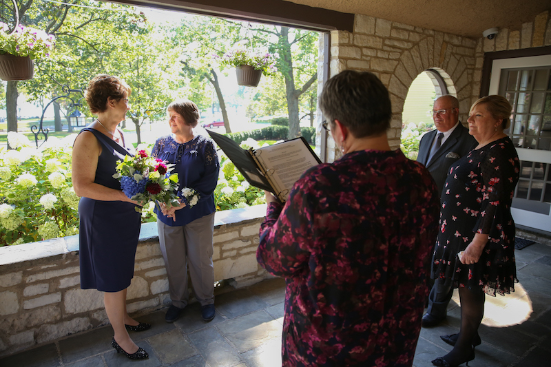 Two women, both wearing navy, stand at the back of a stone porch overlooking trees and greenery. In the foreground, officiant Anita Vaughan conducts the wedding ceremony. Two wedding guests stand and observe.