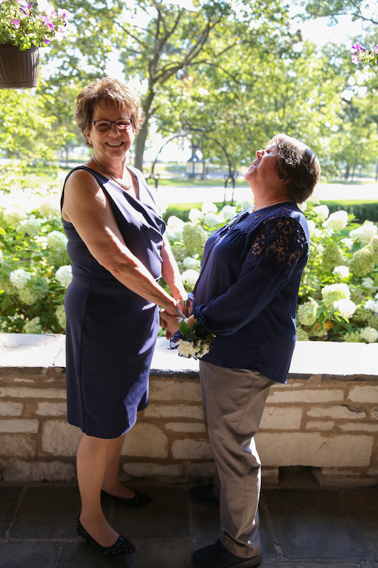 Mary, in a navy dress, and Pam, in a navy short-sleeved top and khaki trousers, stand in front of a low stone wall holding each other's hands during their wedding ceremony.