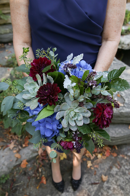 A woman wearing a navy dress holds a bouquet of flowers with bold blue and burgundy flowers and several shades of greenery.