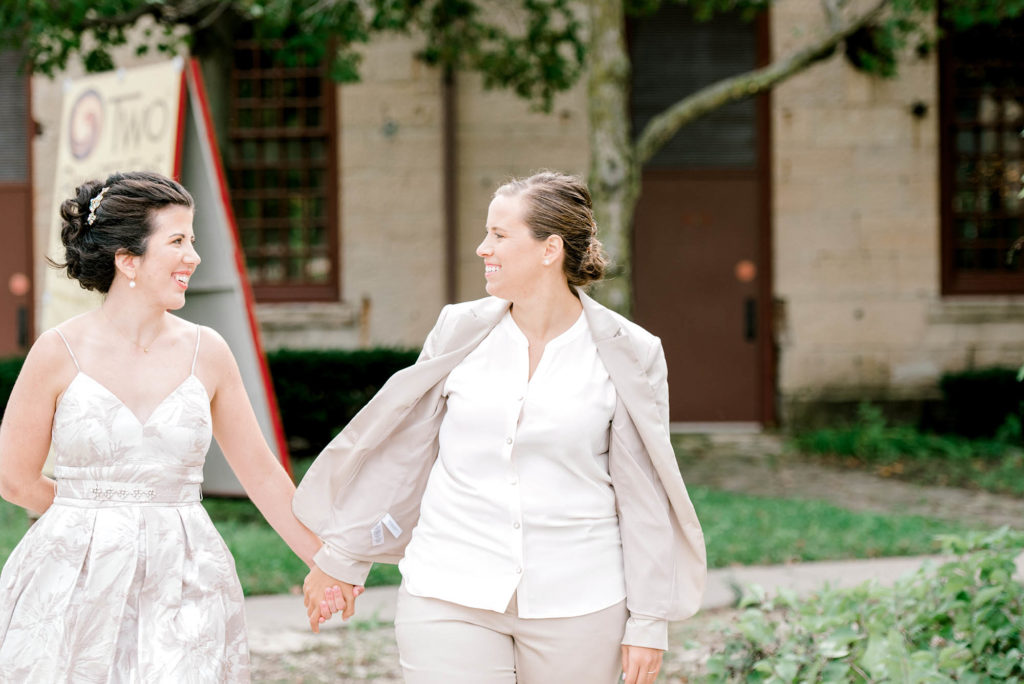 Bride in dress and Bride in suit hold hands with each other