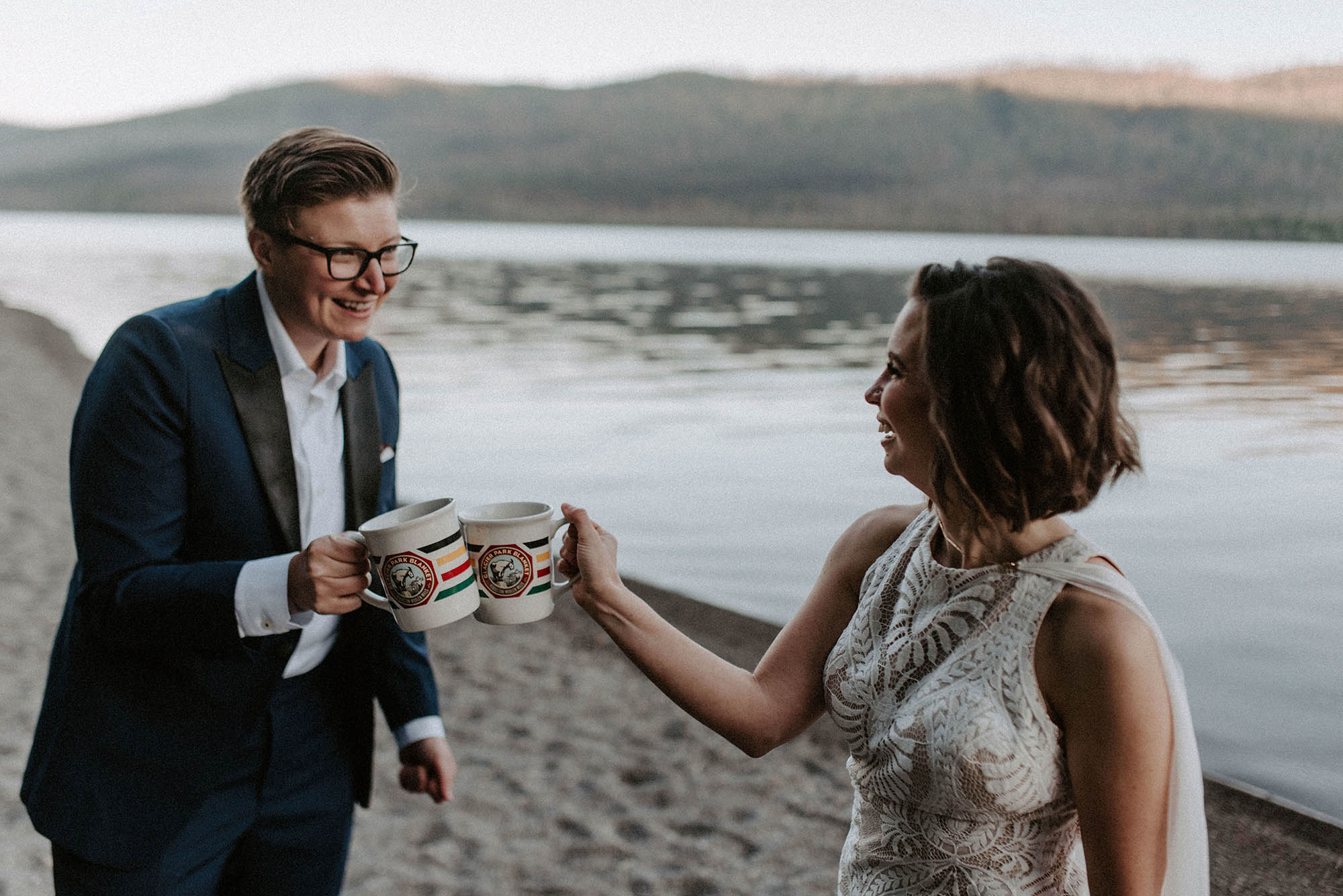 bride in tux and bride in wedding gown click coffee mugs at sunrise on Lake McDonald