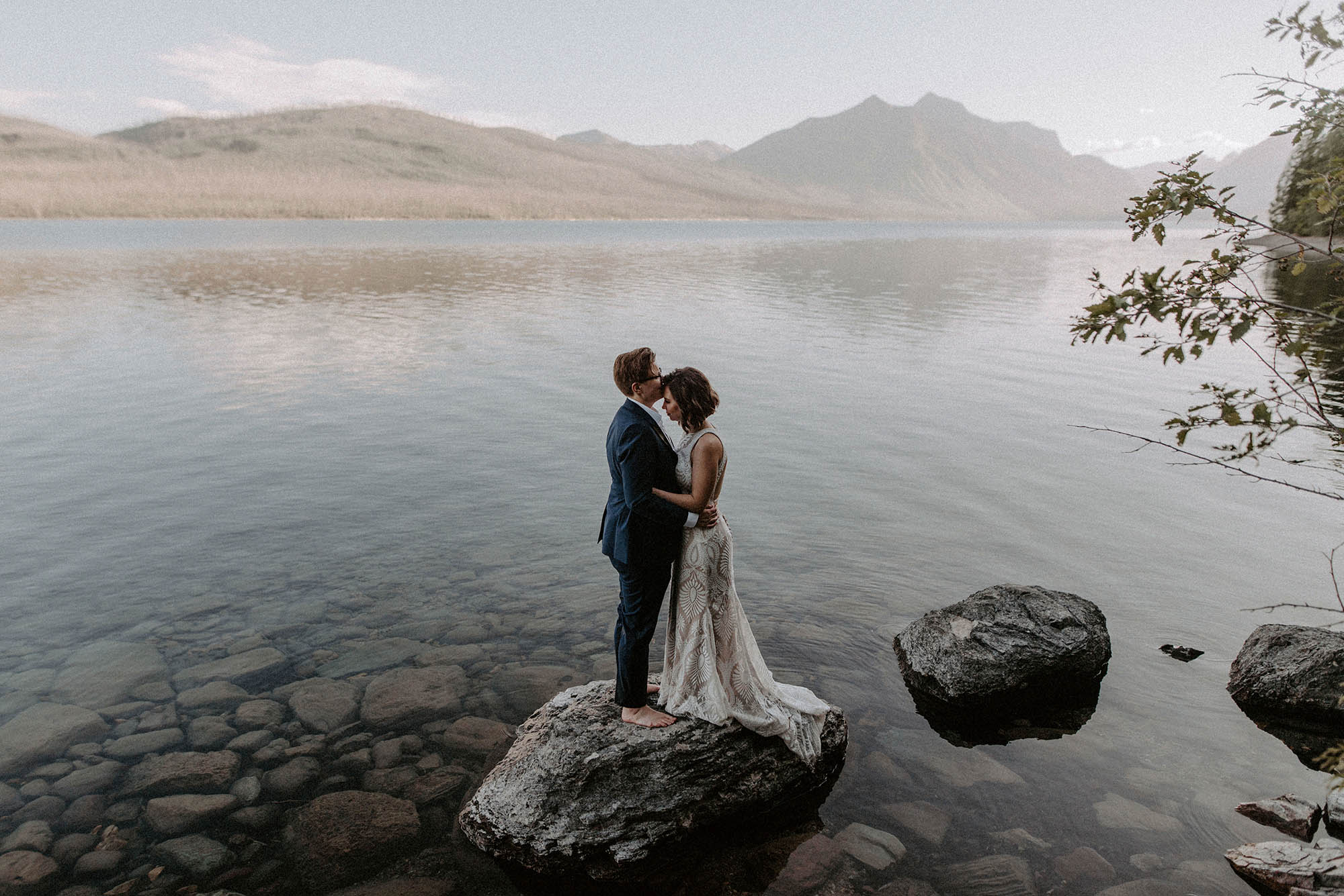 two brides standing on rock in Lake McDonald with mountains behind them