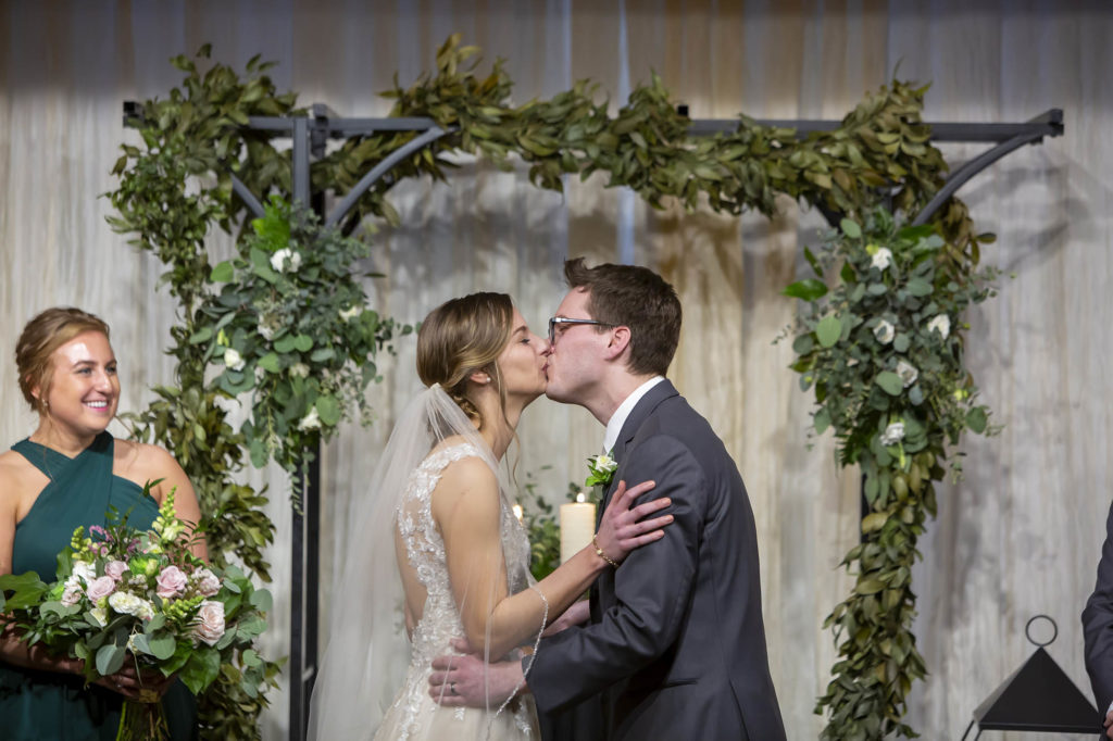 bride and groom kiss at end of wedding ceremony in front of floral arch at Madison Event Center