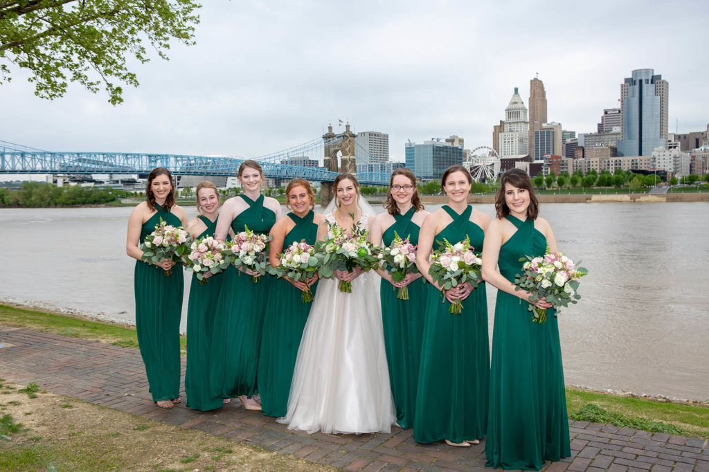 bride and with bridesmaids in green dresses holding pink and green bouquets at Cincinnati skyline