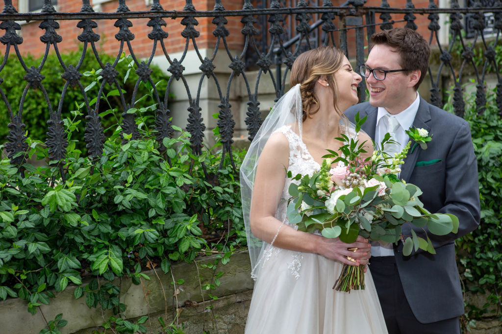 bride in sleeveless wedding gown and veil smiles at groom with glasses and green pocket square
