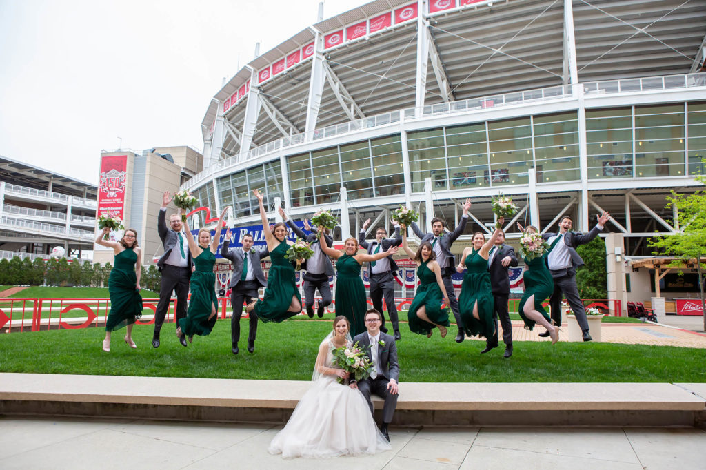 bridesmaids in green dresses groomsmen in gray suits jump up and down at Cincinnati Reds stadium after wedding