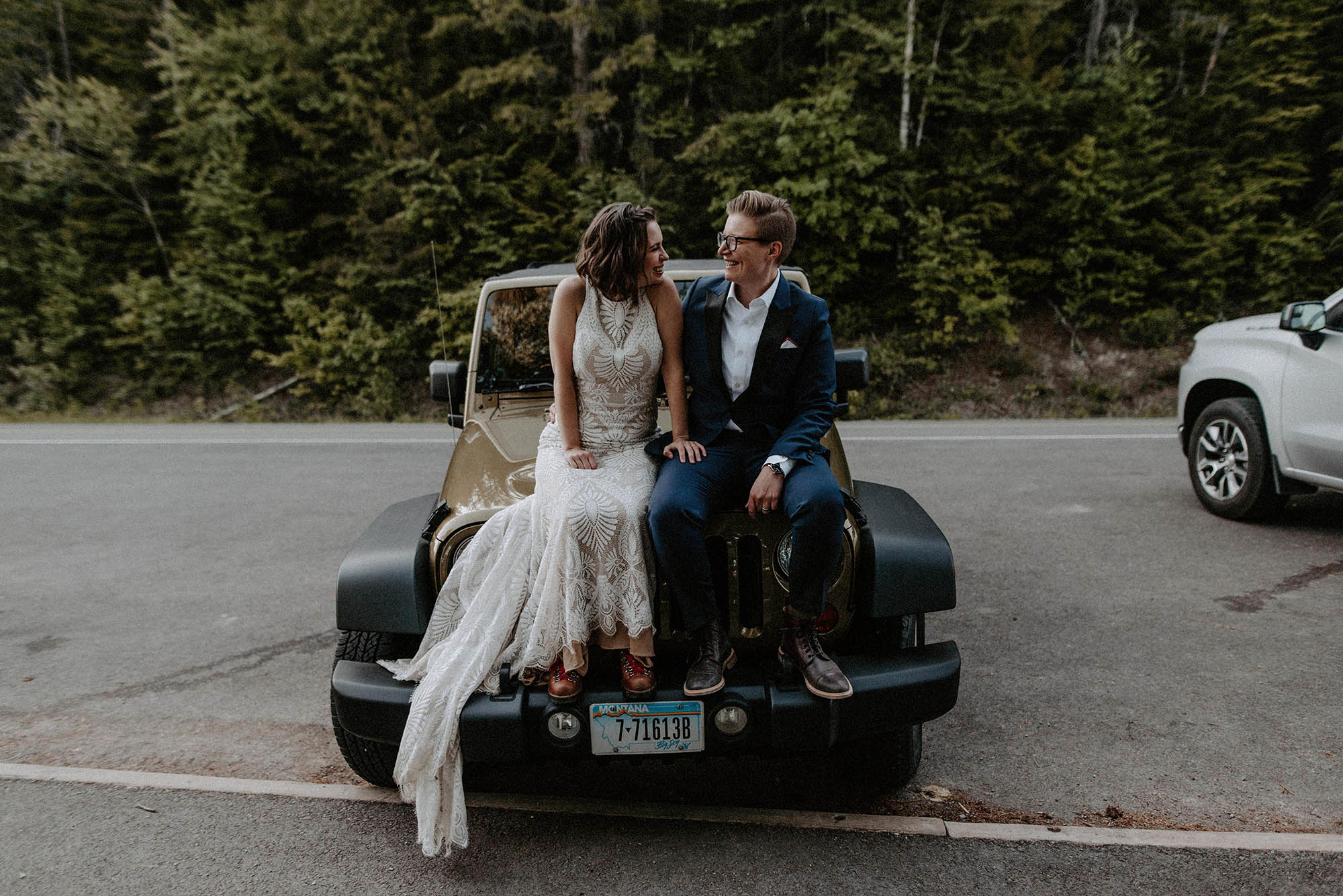 two brides sit on hood of Jeep in front of forest on wedding day