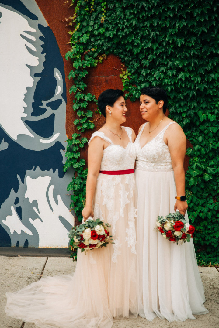 two women in wedding dresses hold bouquets in front of ivy-covered wall