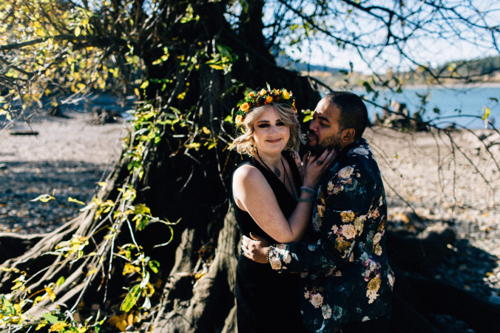 bride in black dress and flower crown smiles while groom in floral jacket embraces her