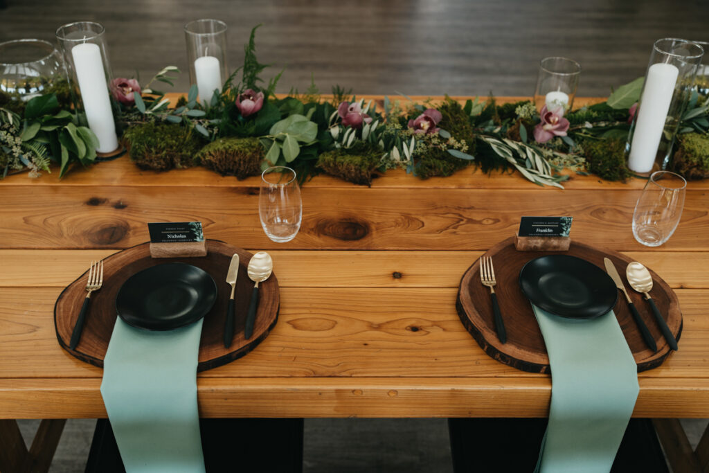 black plates with black and gold flatware on wood chargers on wood farm tables at Trinity Tree Farm wedding. A greenery garland with deep pink orchids is behind them.