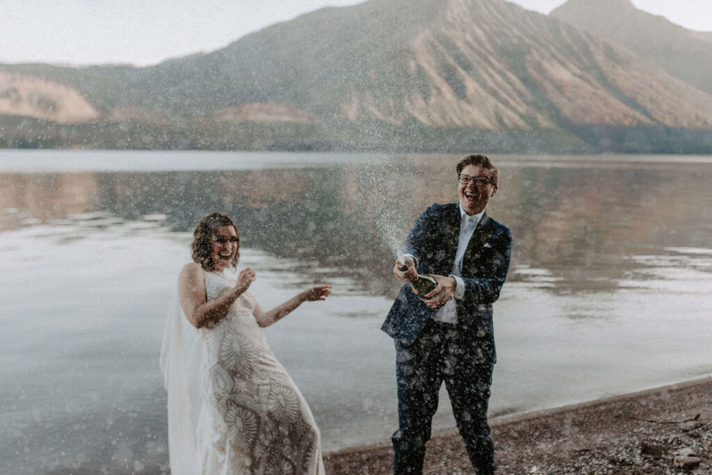 queer couple popping champagne next to lake at their wedding