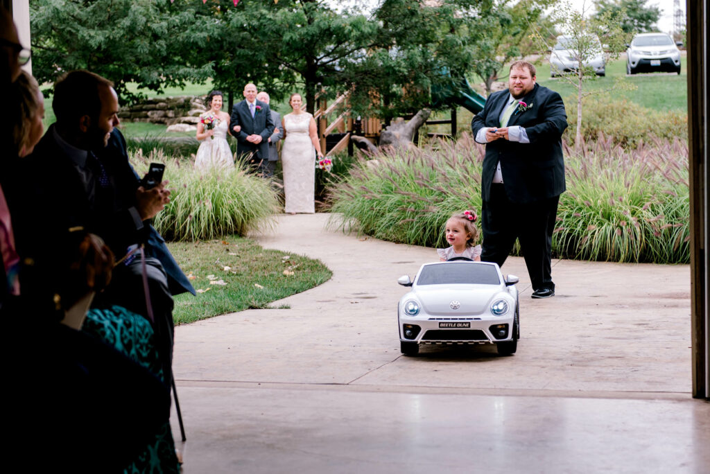 man in suit standing behind little girl driving toy car