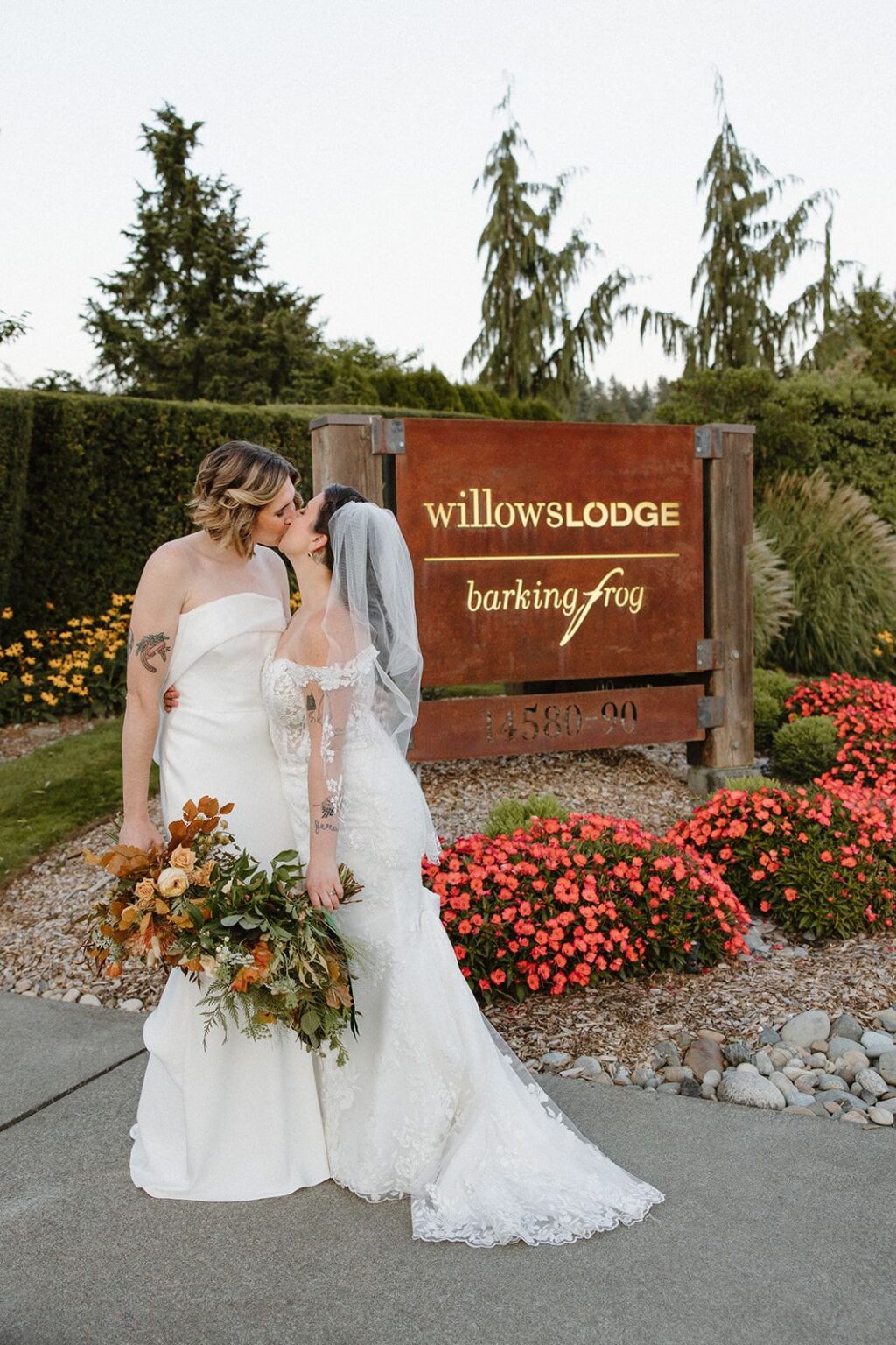 Two brides kiss in front of Willows Lodge wedding venue sign.