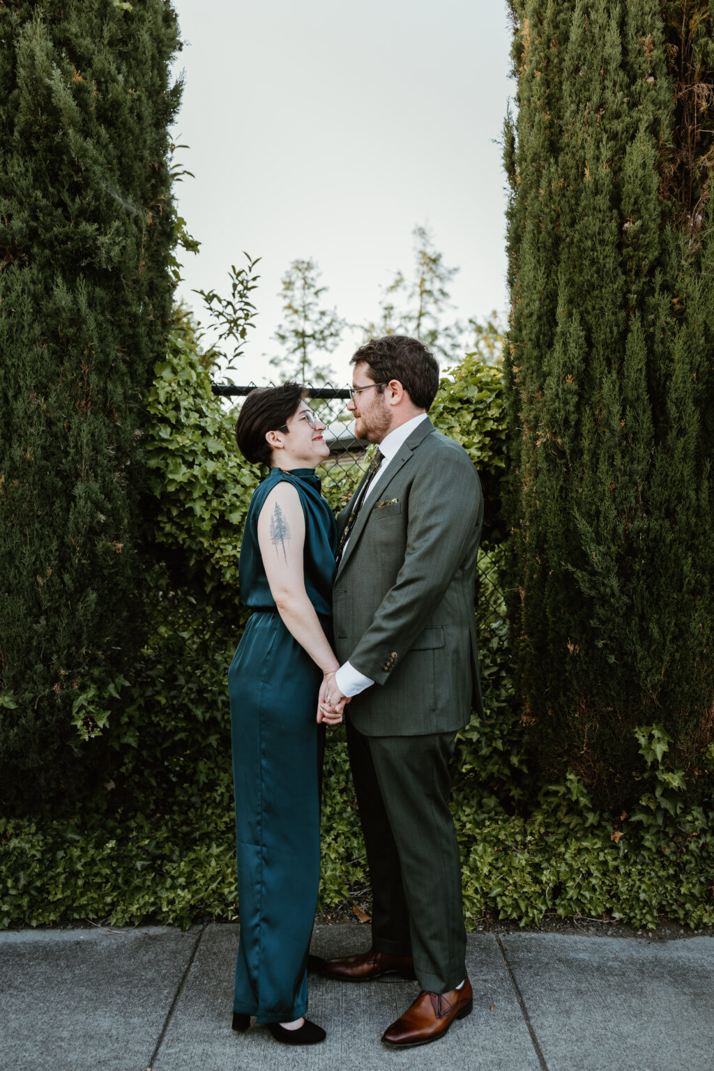 non-binary marrier in teal jumpsuit with groom in gray suit in front of mossy trees