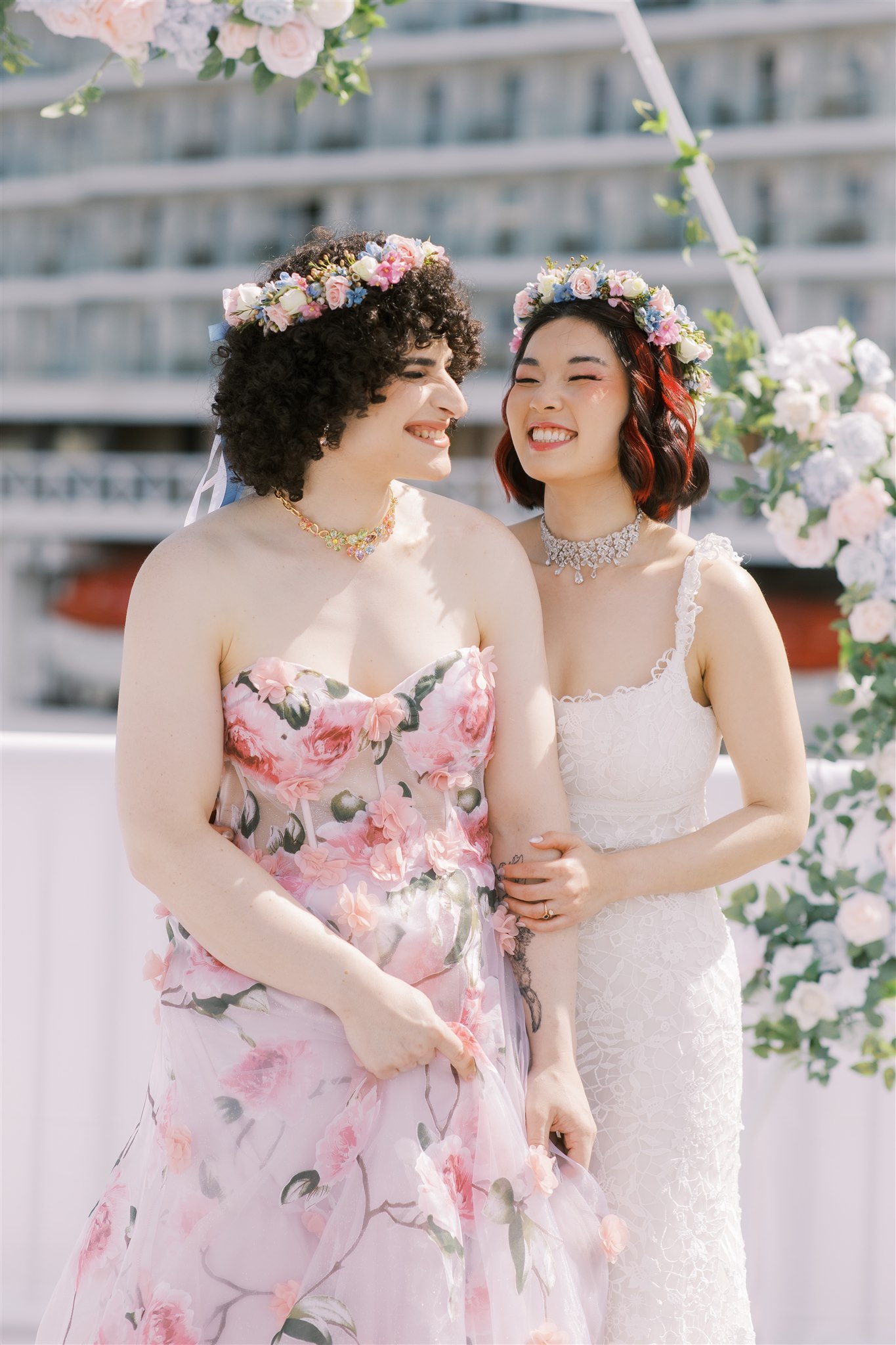 bride in pink floral gown and bride in white gown, both with flower crowns, smile at each other in front of floral arch